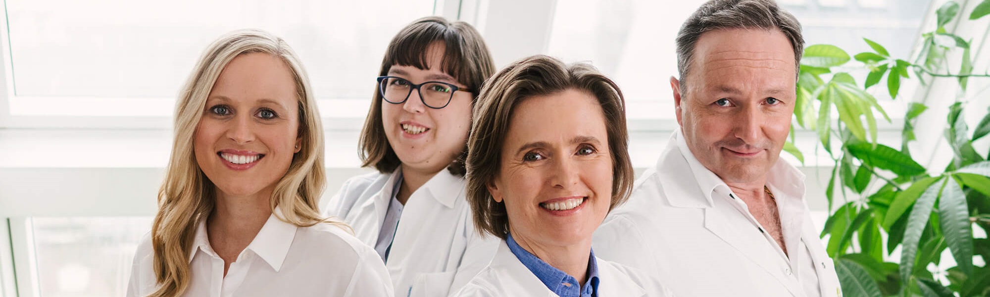 Gruppenfoto mit 3 Frauen und einem Mann rechts die alle weiße Ärztekittel tragen und in die Kamera lächeln.